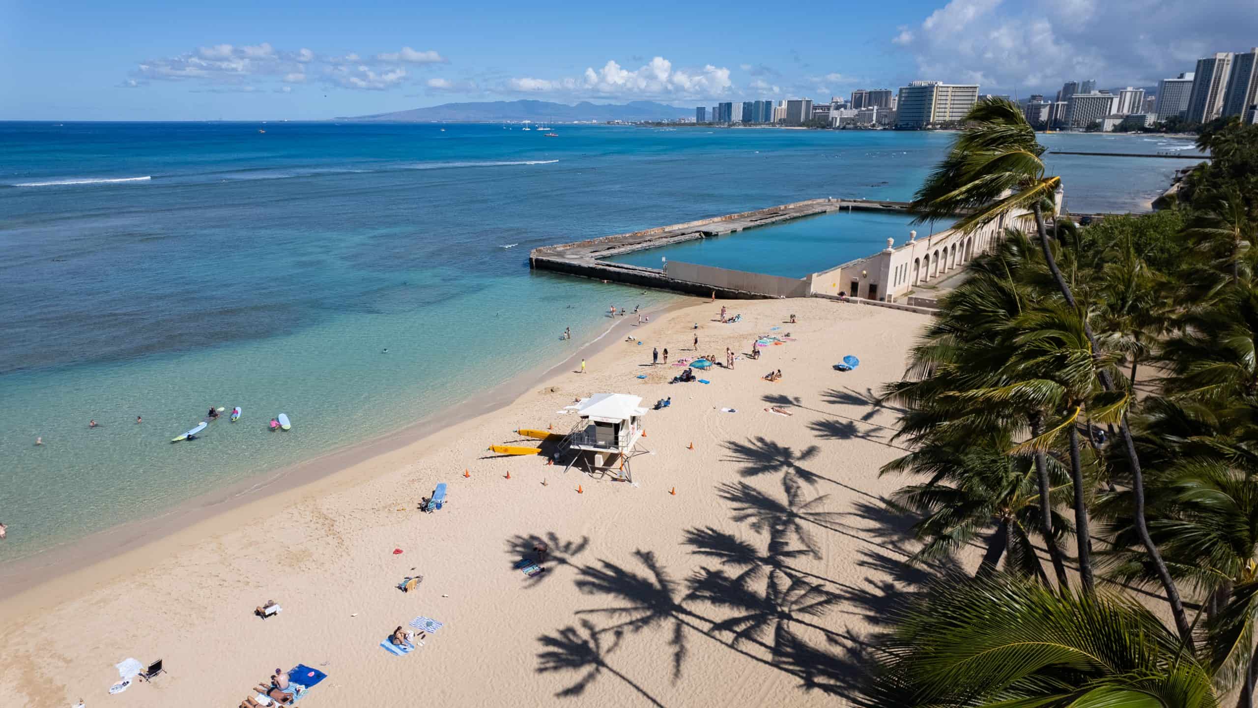 An aerial view of Kaimana Beach with people on it and palm tree shadows, with the Natatorium War Memorial and Waikiki in the background