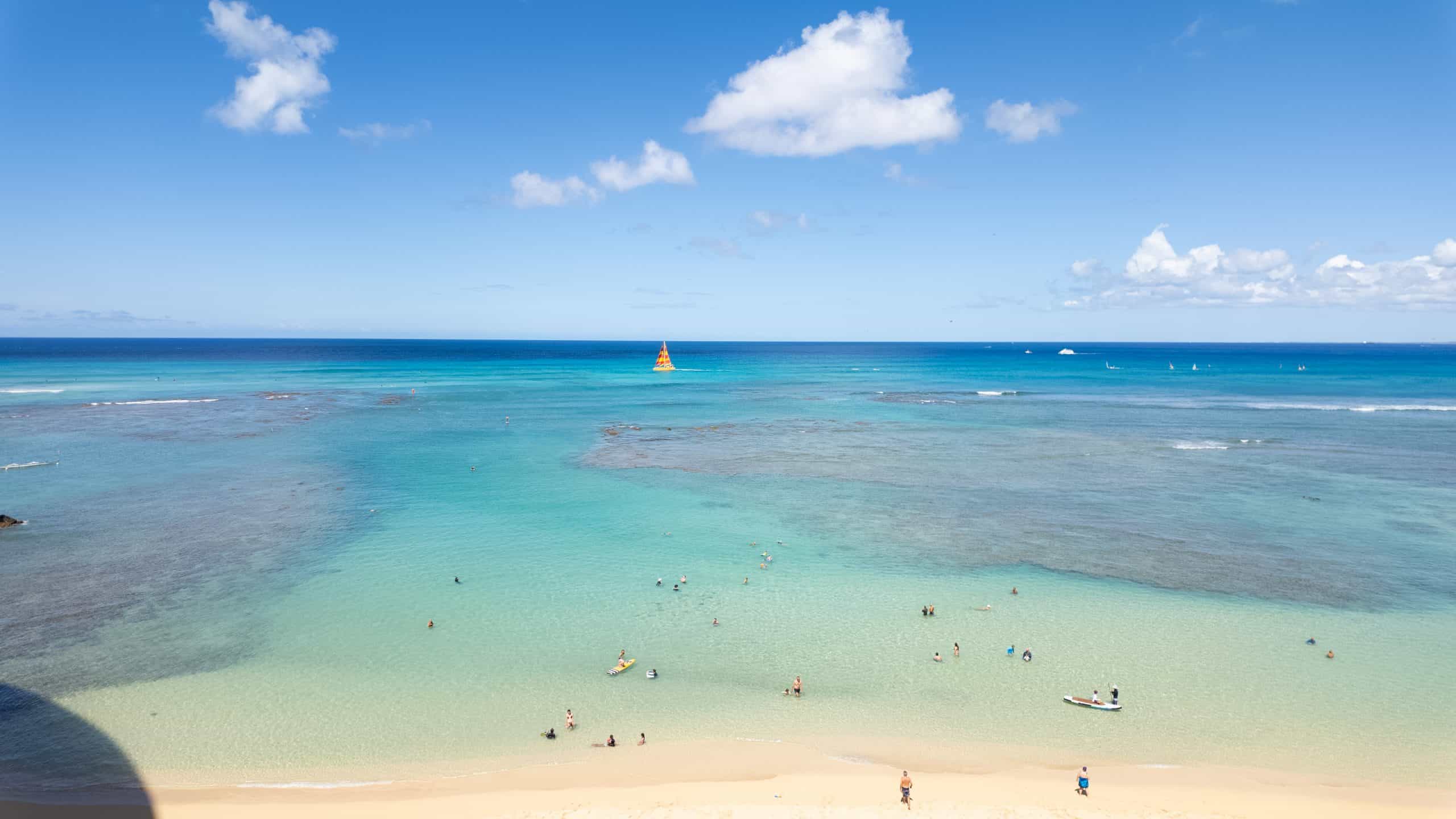 People in the water in a clear water sunny beach with a red and yellow sailboat in the background