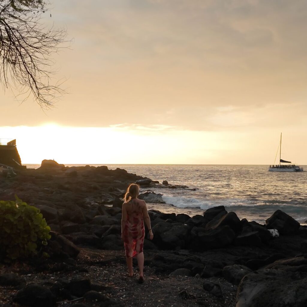 A blonde woman standing on lava rocks watching a sunset with a sailboat in the background