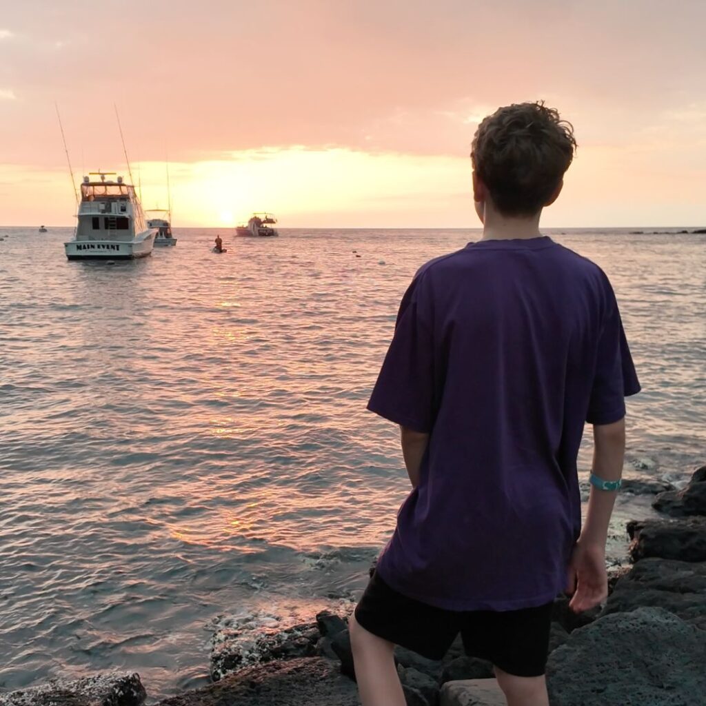 A young man standing on rocks watching a sunset over an ocean with boats in the distance