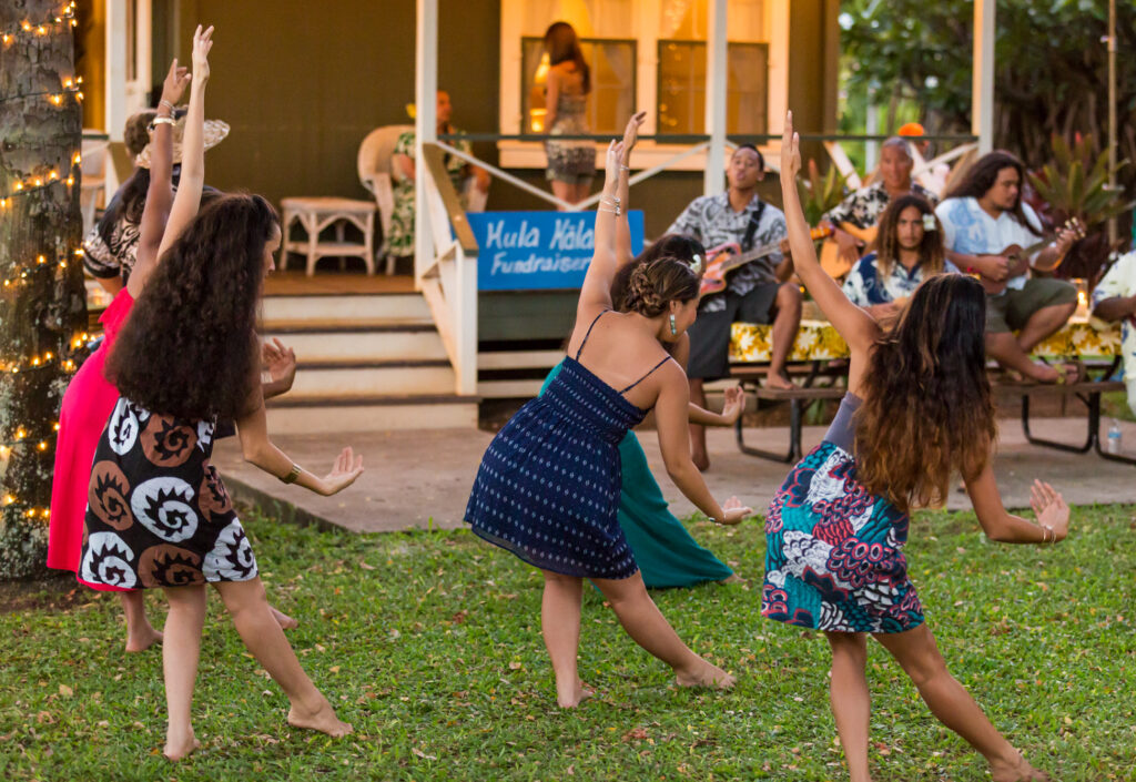 5 women in colorful shirts and dresses dancing hula with 4 musicians playing guitar in the background