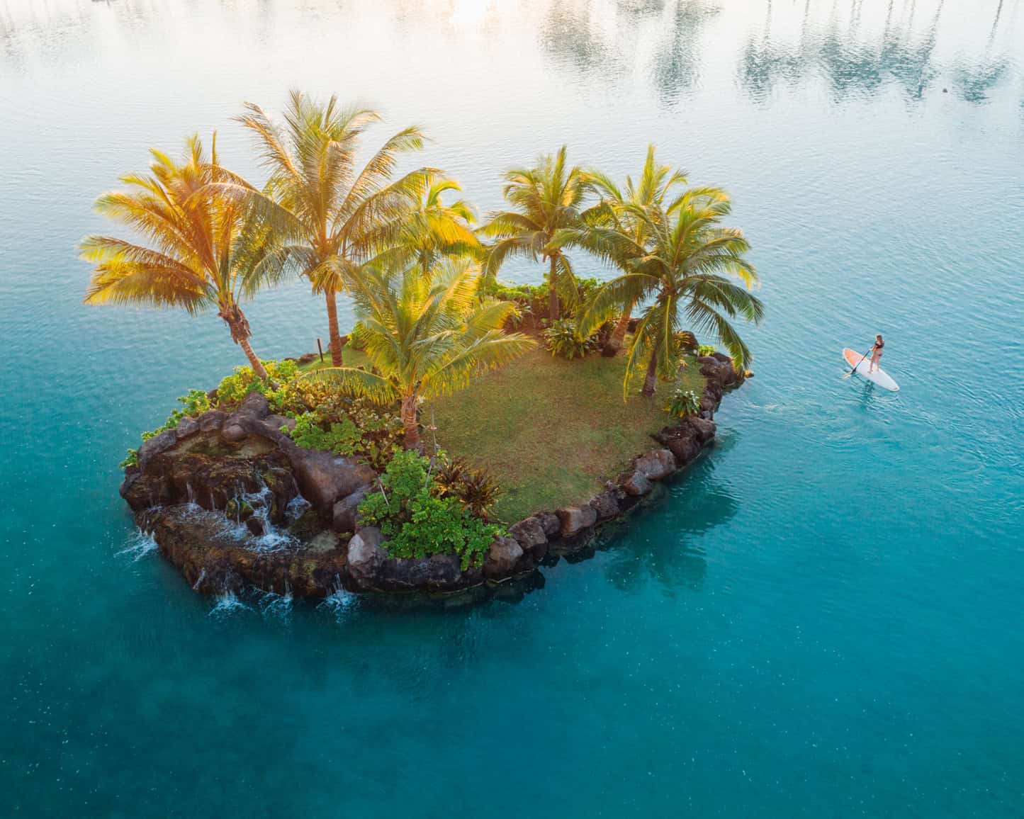 A woman in calm ocean water on a paddle board near a small island with palm trees on it.