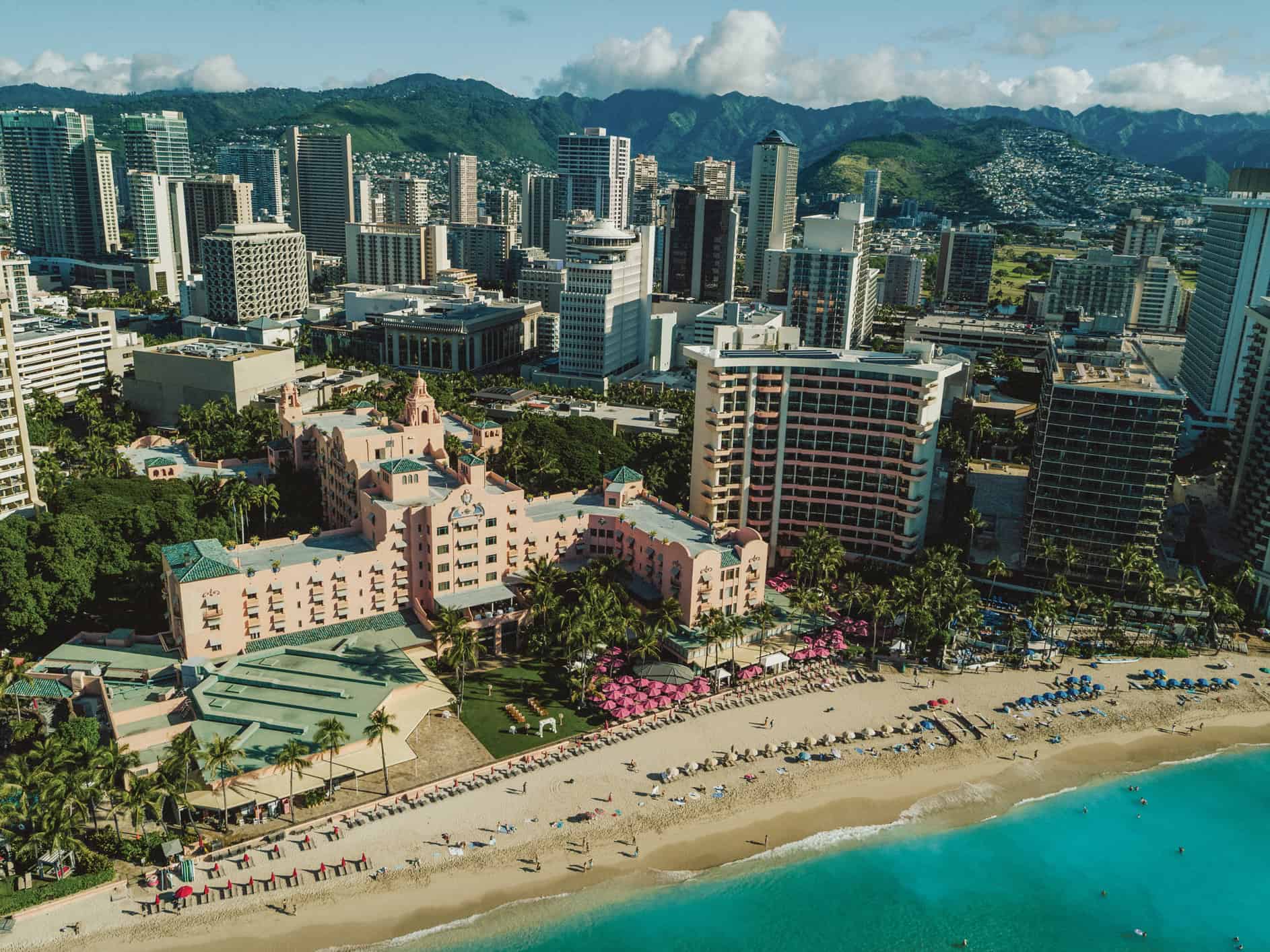 An aerial view of a pink building with people on the sand in front of it and a city with mountains in the background