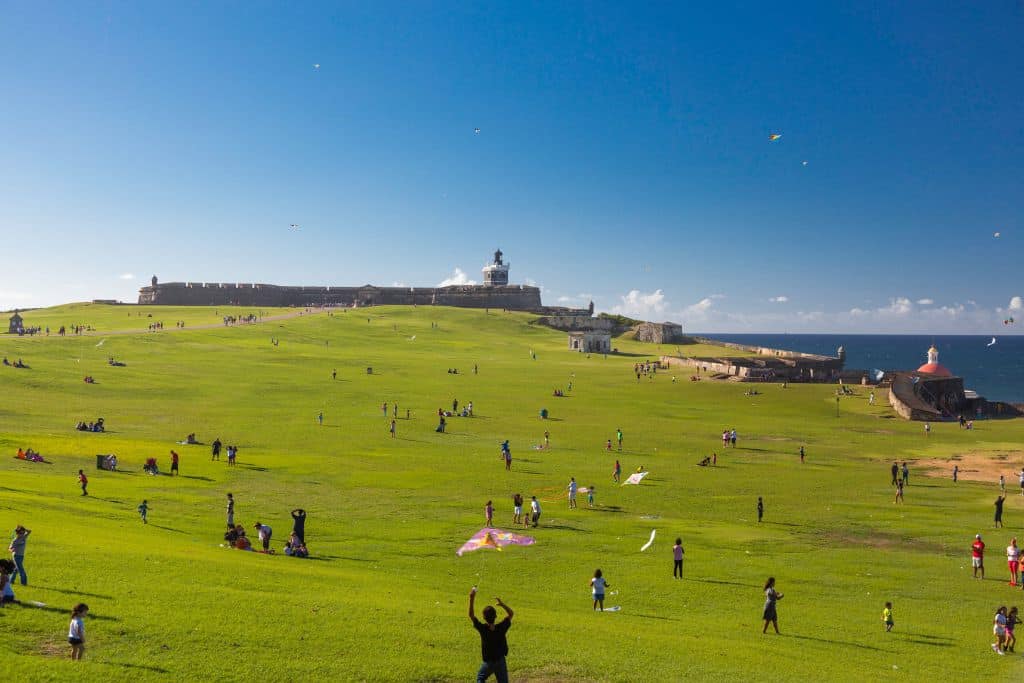A large green lawn with an ancient building in the background and the ocean to the right. The people on the lawn are hanging out and some are flying kites.