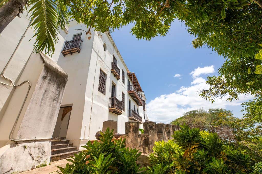 An old white building with some windows and balconies and weathering on the side of it, surrounded by an old looking stone fence and brightly green colored tropical trees.