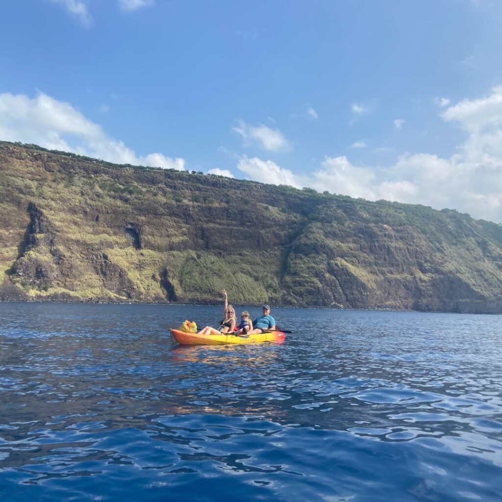 3 people in a bright yellow kayak with their hands up smiling and cliffs behind them