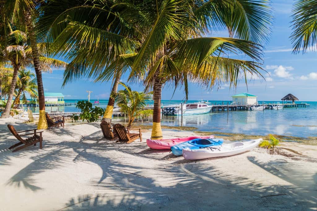 White sand with palm tree shadows, brightly colored boats and lounge chairs lining the edge of the calm water with palm trees scattered about and a dock with sailboats in the background.