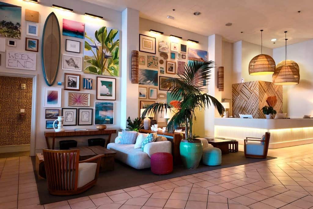 Lobby of Kaimana Beach Hotel with a tropical gallery wall, rattan pendant lights, and cozy seating areas with coastal-style furniture.