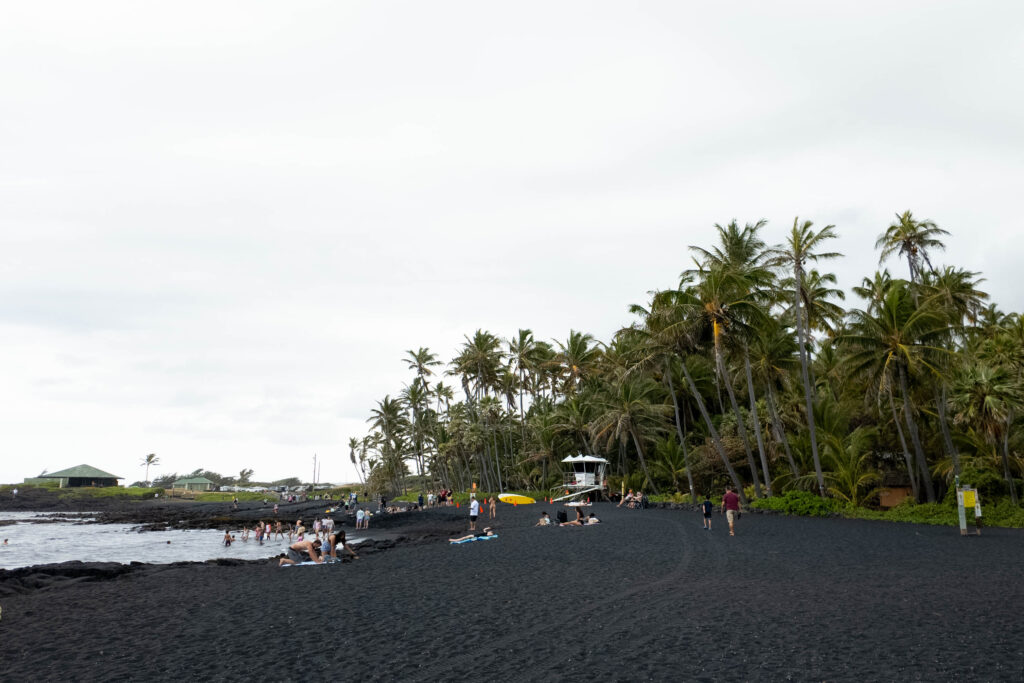 A black sand beach with people on it, a lifeguard, yellow surfboard and palm trees in the background