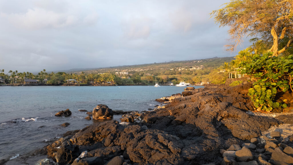 A bay with lava rocks in front and lush tropical plants in the background, and boats in the distance