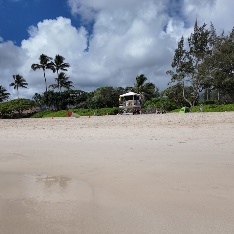 A large stretch of white sand with a lighthouse and palm trees in the distance