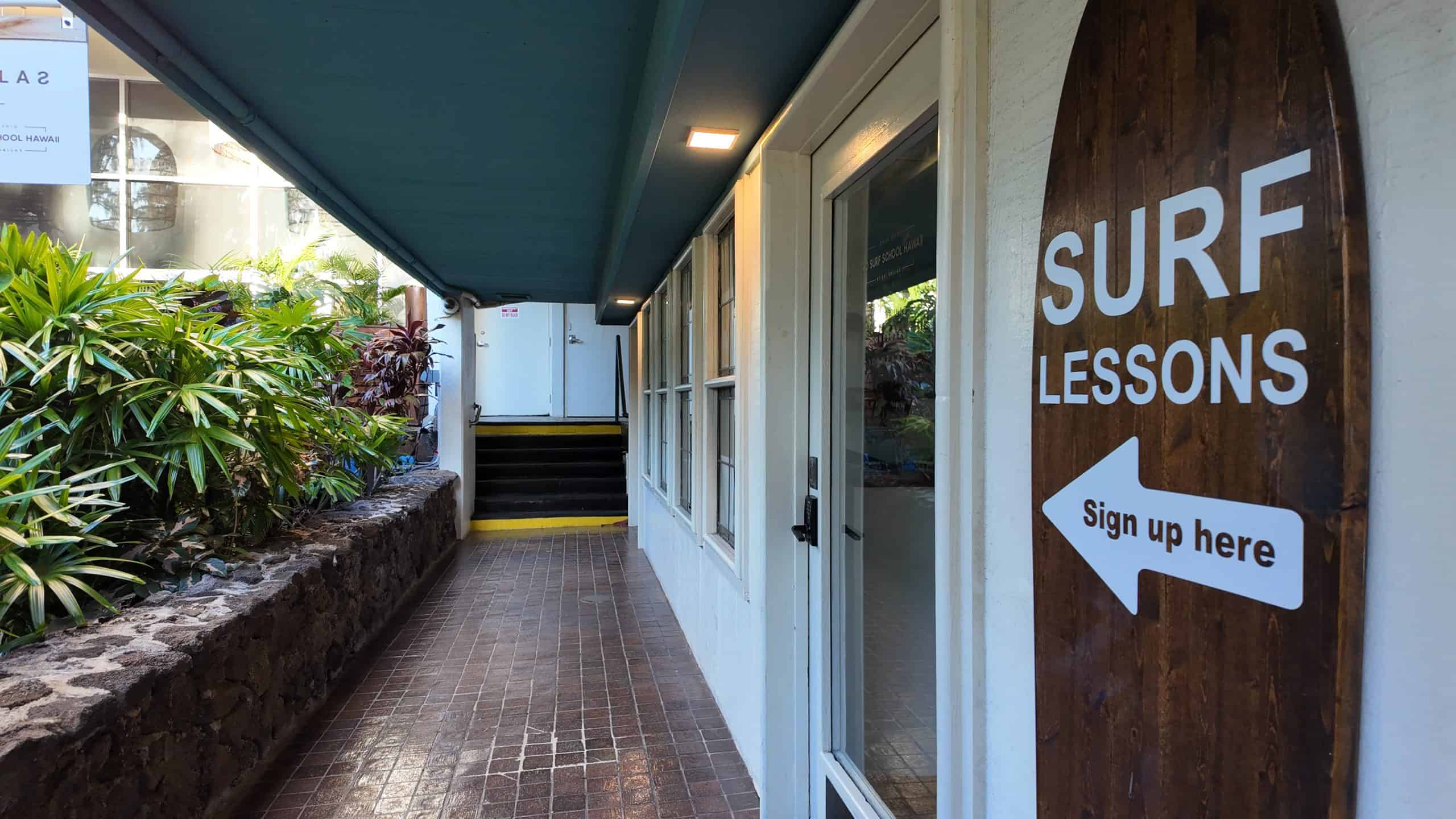 Wooden surfboard-shaped sign for surf lessons at the entrance to a walkway at Kaimana Beach Hotel.