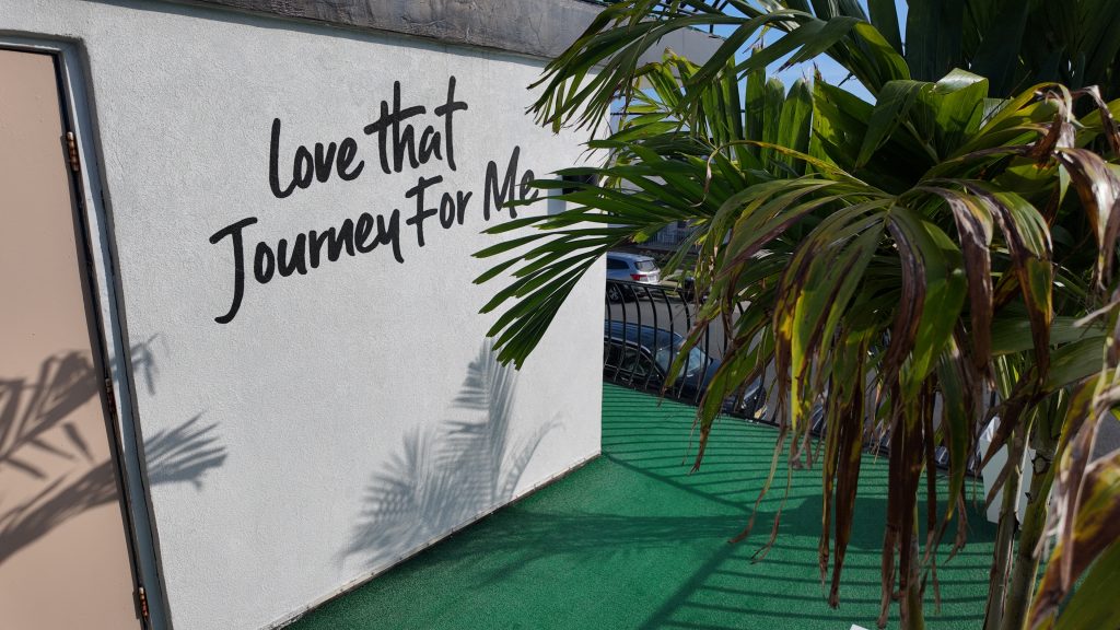 A hotel wall that says "love that journey for me" with tropical trees around it