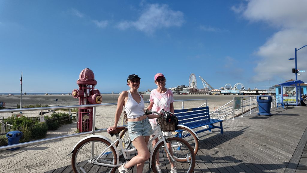 A blonde woman in a white top and black hat and a woman in a pink shirt and pink hat on the boardwalk with bikes in Wildwood NJ
