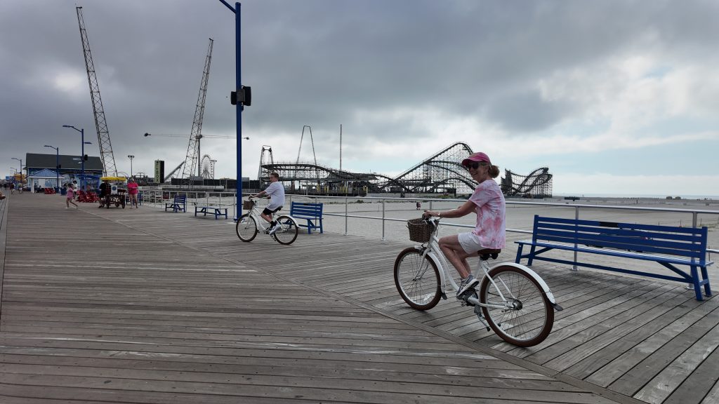 A woman and a teenager biking on a boardwalk in front of a rollercoaster