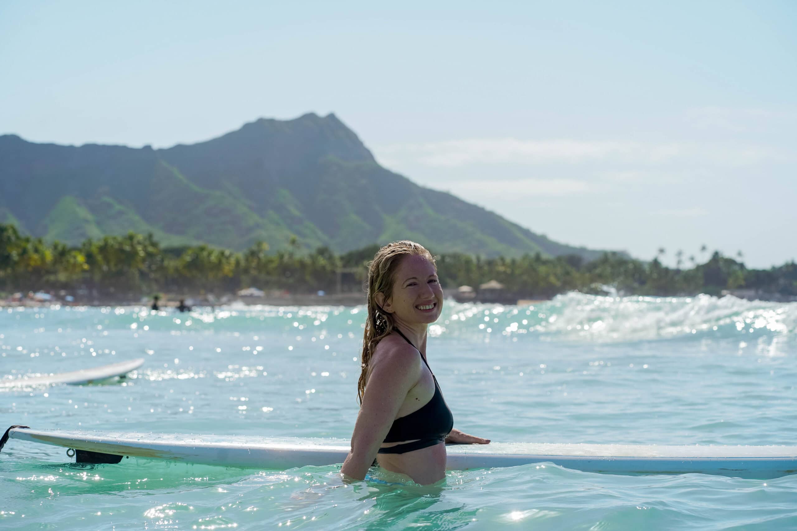 A blonde woman in the water with a surfboard and Waikiki Beach behind her