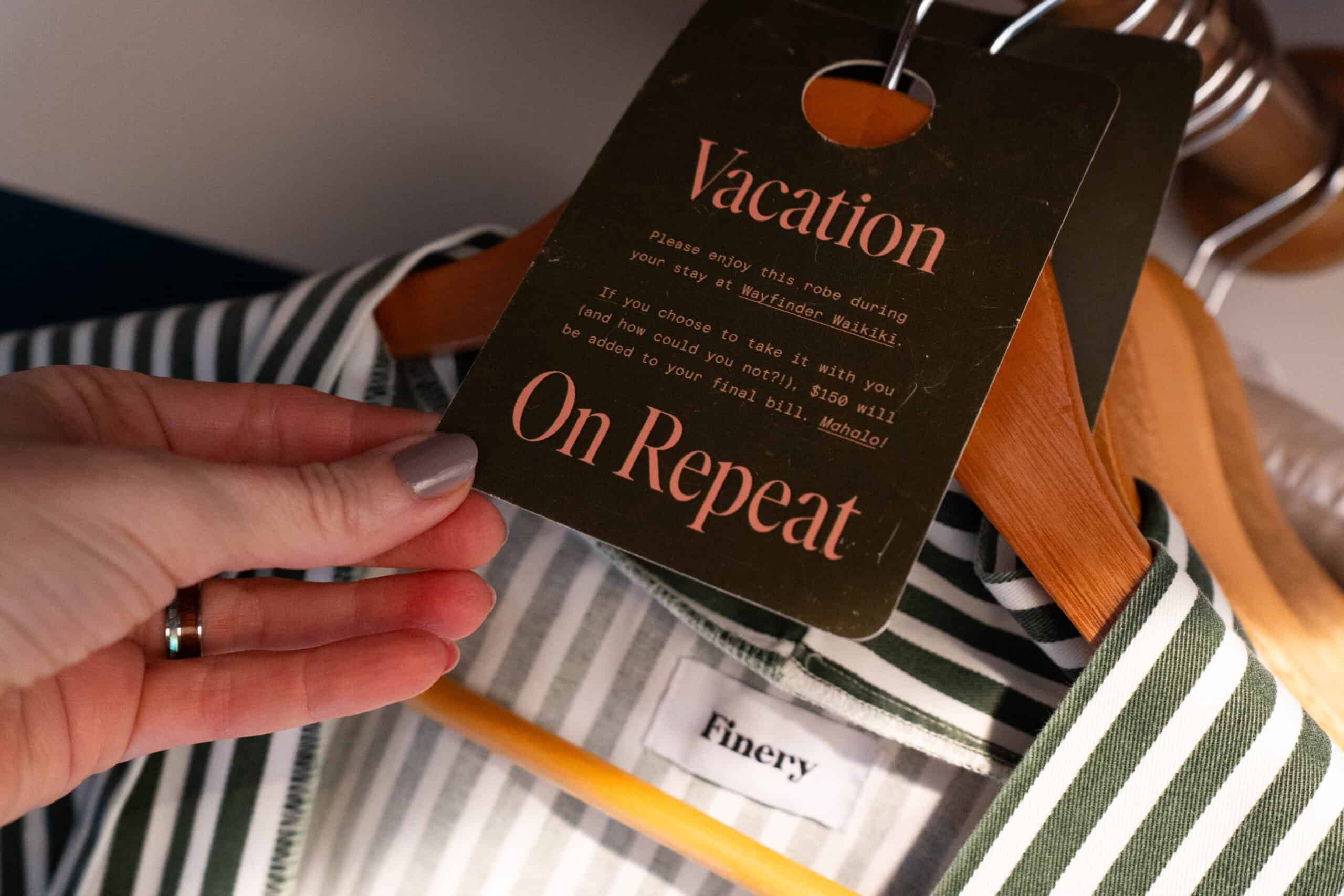 A womans hand with tan painted nails holding a tag that says vacation on repeat hanging on a green and white striped robe.