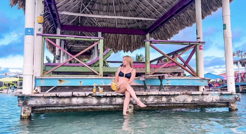 blonde-woman-sitting-on-dock-with-coconut-at-pirates-sectre-beach-amvergris-caye-belize