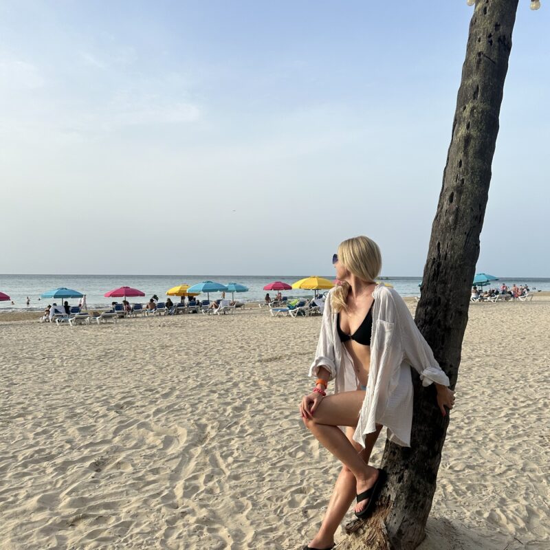 A blonde woman in a white shirt leaning against a palm tree, looking towards the ocean with brightly colored umbrellas in front of it.