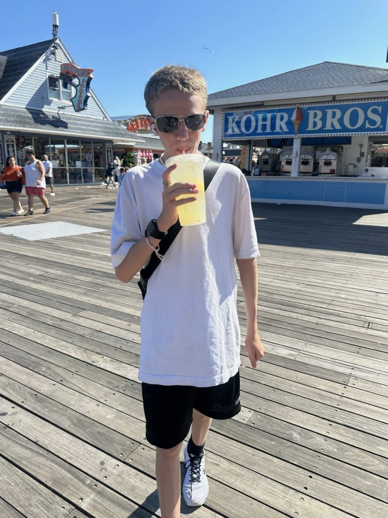 A boy in a white tee shirt and sunglasses drinking lemonade on the boardwalk
