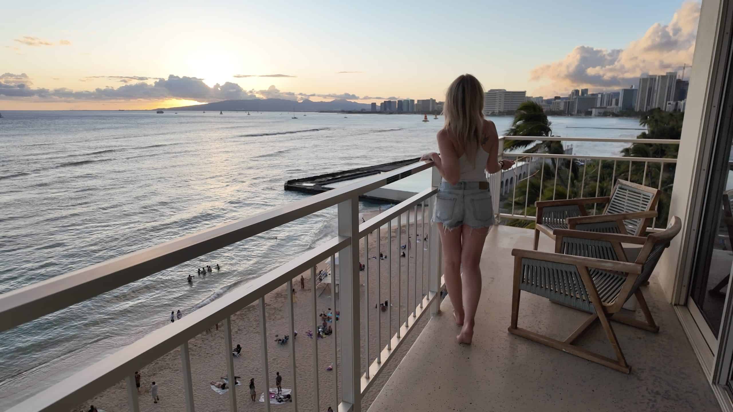 A blonde woman standing on a patio looking towards Waikiki beach at sunset.