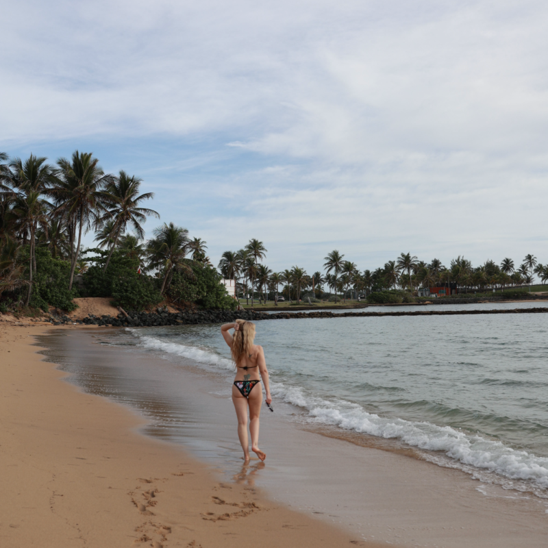 A blonde woman with her hand in her hair walking along the ocean with palm trees in the background.